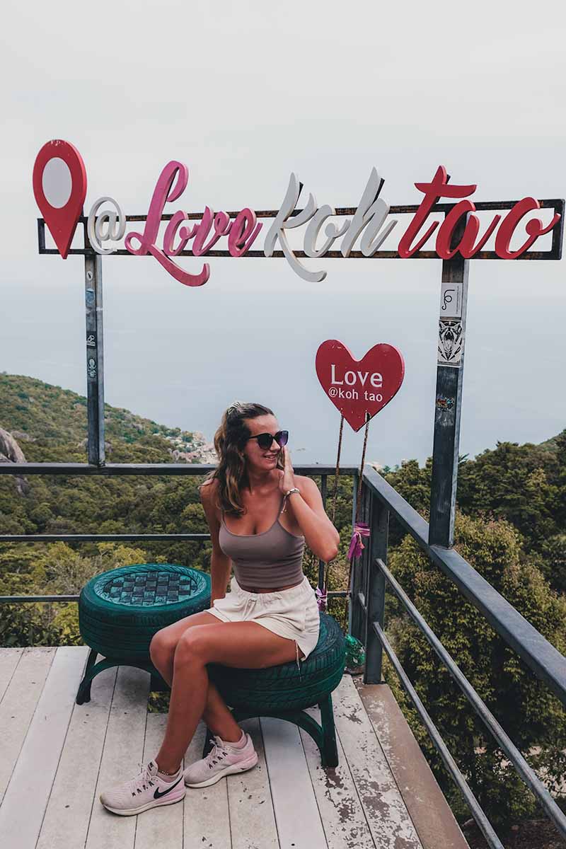 A tourist sitting under the Love Koh Tao sign with the jungle-covered east coast of Koh Tao in the background