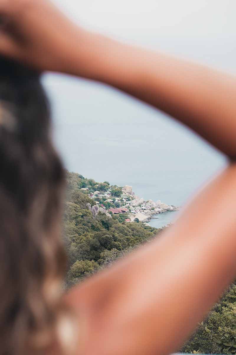 A tourist with a hand on her head at the Love Koh Tao Viewpoint with the jungle-covered east coast of Koh Tao in the background