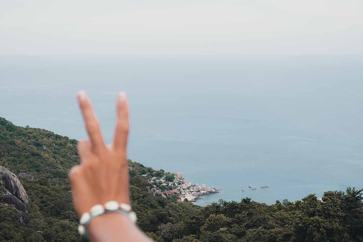 A tourist's hand displaying the peace sign at the Love Koh Tao Viewpoint with the jungle-covered east coast of Koh Tao in the background