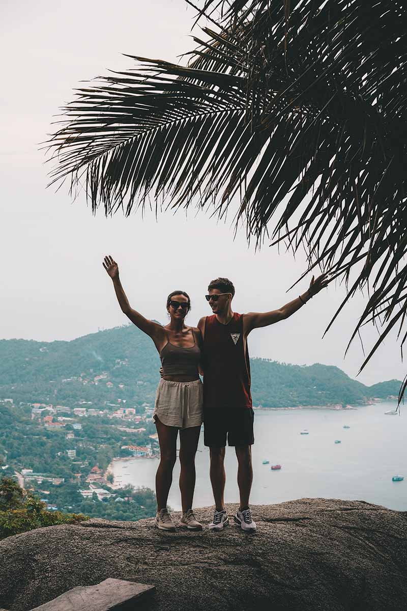 Two tourists standing with raised arms at Summit Viewpoint with the west coast beaches and jungle-covered south of Koh Tao behind them