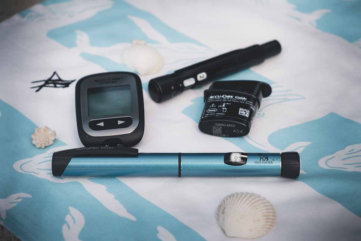 A blue Novorapid insulin pen, Accu Chek blood glucose meter, box of blood test strips, lancet and white shells laid out on a turquoise and white beachtowel