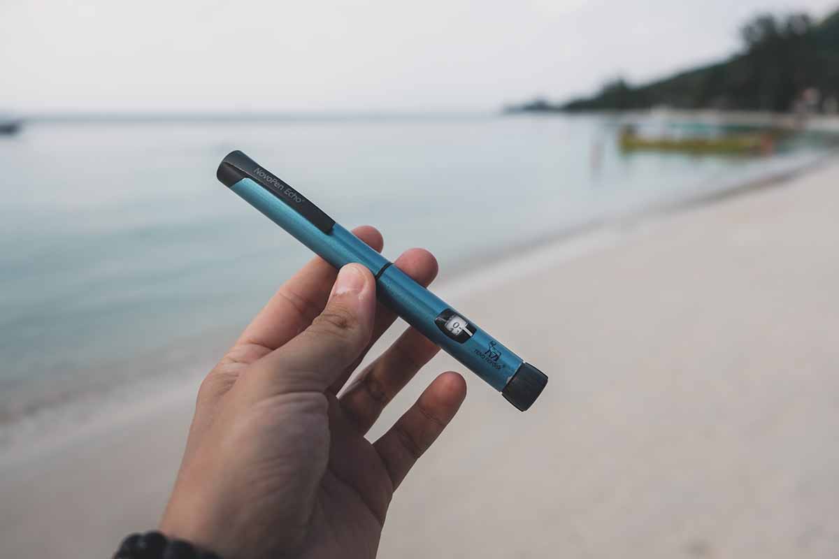 A hand holding a blue Novorapid insulin pen with a white sand Thai beach in the backgroumd