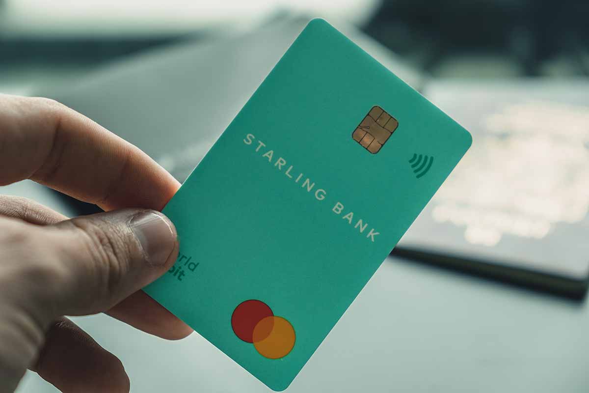 A hand holding a green Starling Bank debit card with a blurry United Kingdom passport in the blurred background