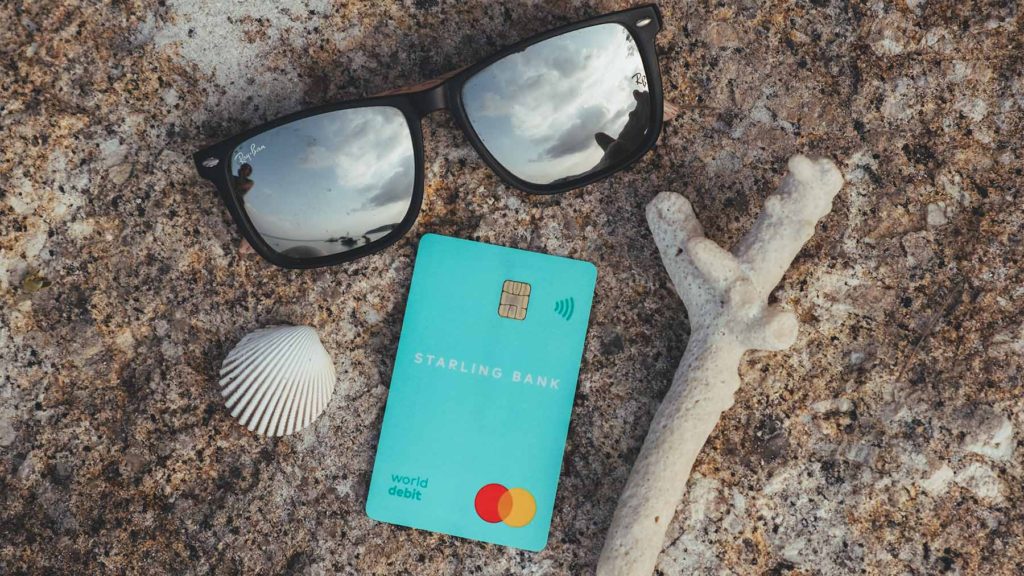 A green Starling Bank debit card, two white shells and a pair of sunglasses with reflective lenses laid out on a rock