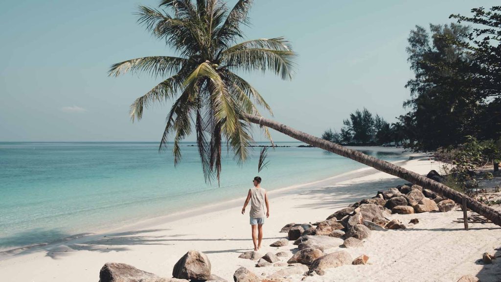 A tourist walking on Chaloklum Beach next to rocks and an overhanging palm tree.