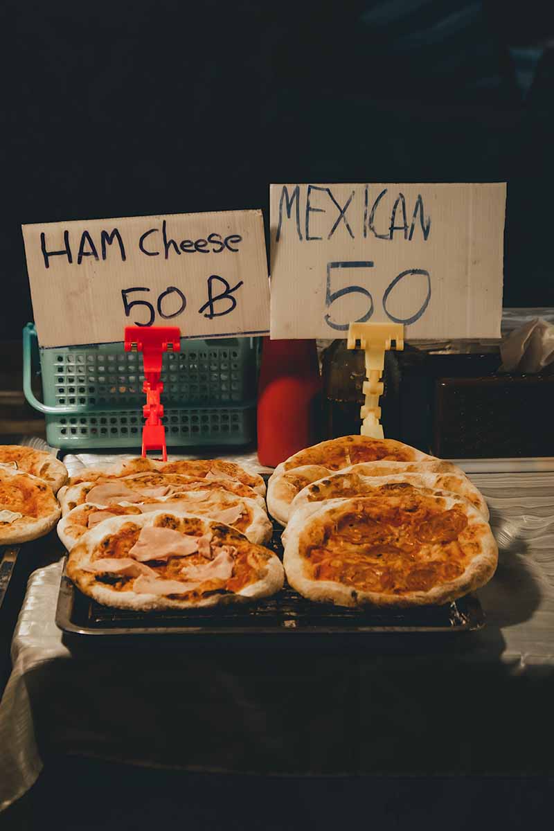 A market stall displaying ham and cheese, and Mexican pizzas below handwritten, cardboard signs advertising them for 50 baht.