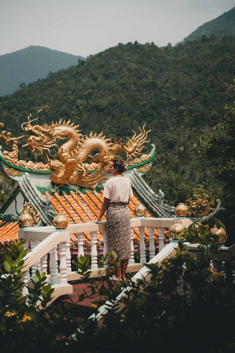 A tourist standing on the temple's upper level landing with layers of the surrounding mountains in the background.