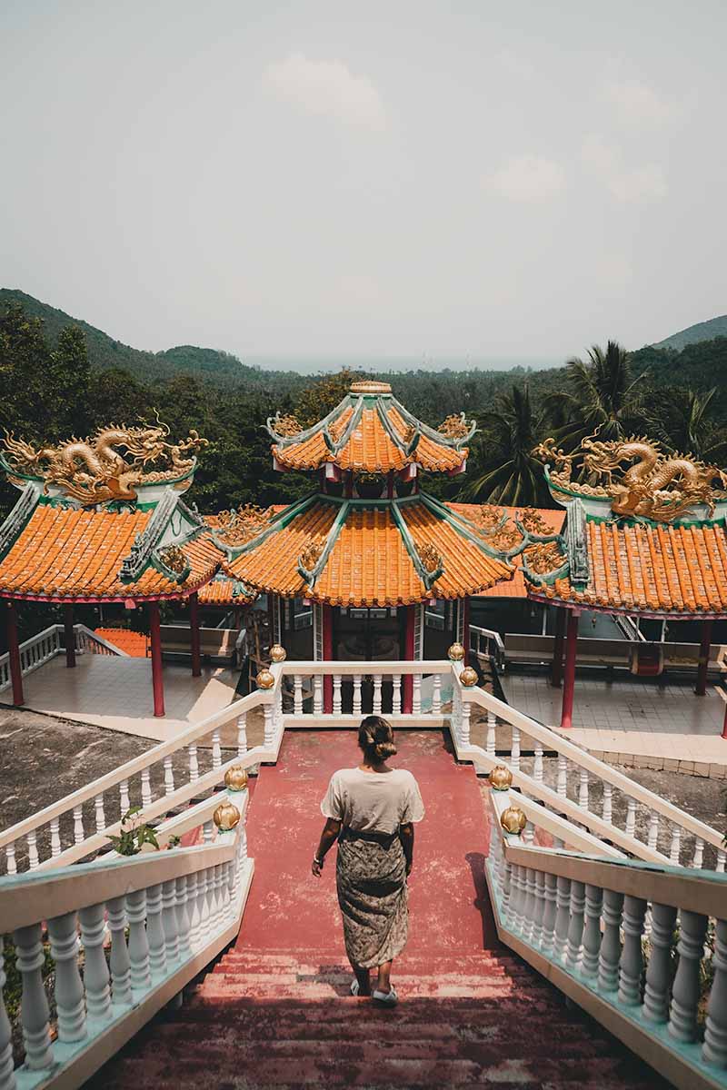 A tourist walking down the stairs of the temple's upper level which looks over the bright orange and teal roof of the lower level, mountainous surrounding, and distant coastline.