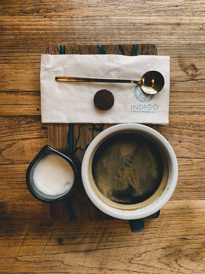 An americano coffee, small jug of milk, biscuit, gold teaspoon and napkin served on a wooden tray served by Indigo cafe.