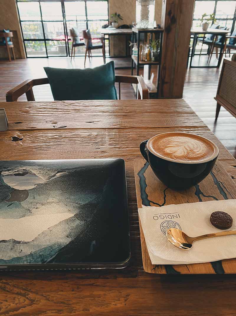 A wooden table with a laptop and wooden tray with a cappuccino, biscuit, gold teaspoon and napkin in Indigo cafe's upstairs seating area.
