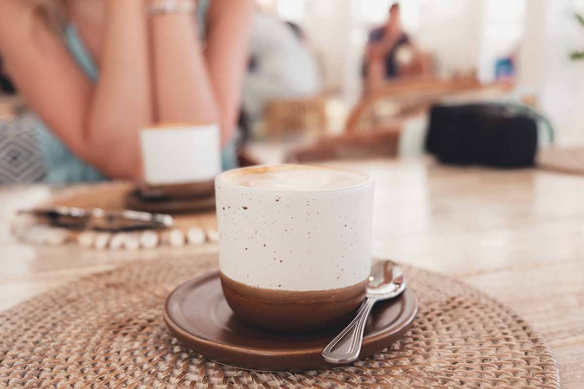 Cappuccino in a white and brown mug on a cafe table with Sand and Tan restaurant's interior blurred in the background.