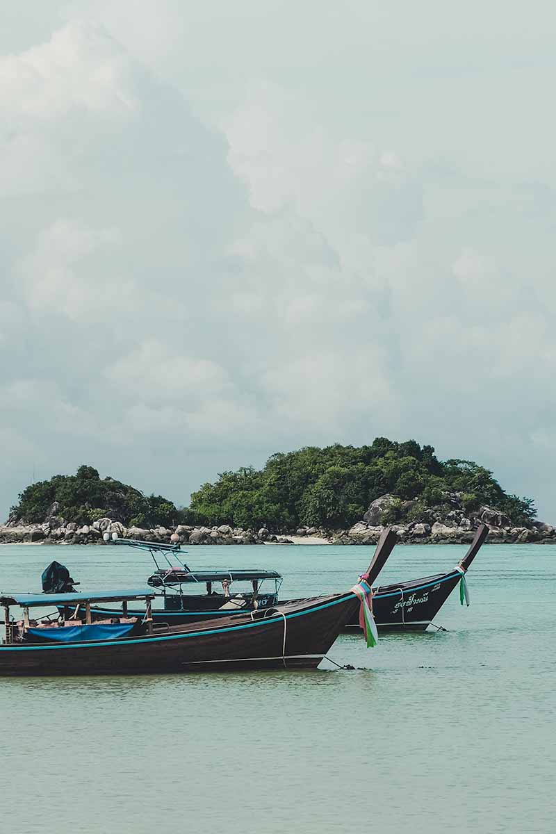 Two long-tail boats anchored in the water with the tiny island of Koh Kra in the background.