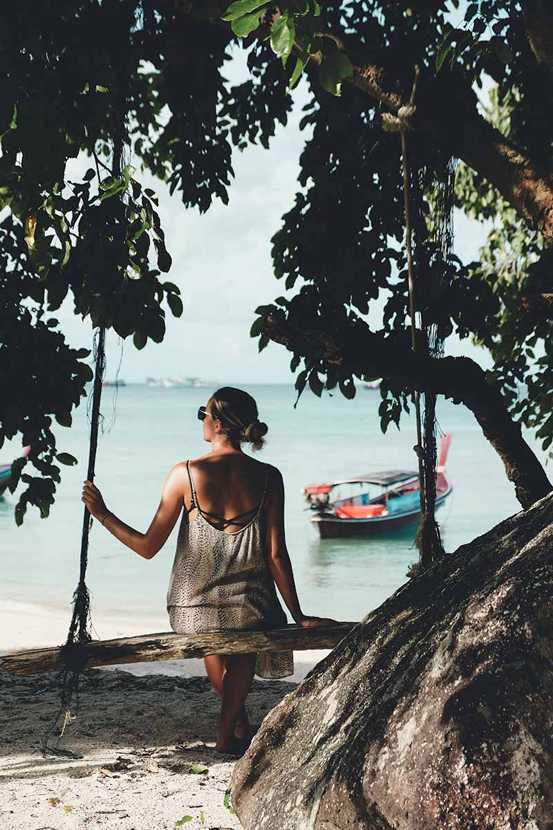 A female tourist sitting on a rustic wooden swing under a tree on the beach.