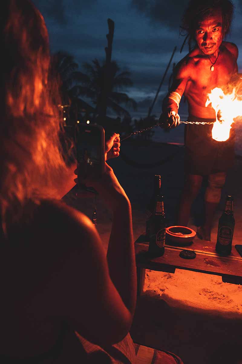 A fire performer holds one end of his fire ball chain while a sitting tourist holds the other end with one hand and films on their phone with the other.