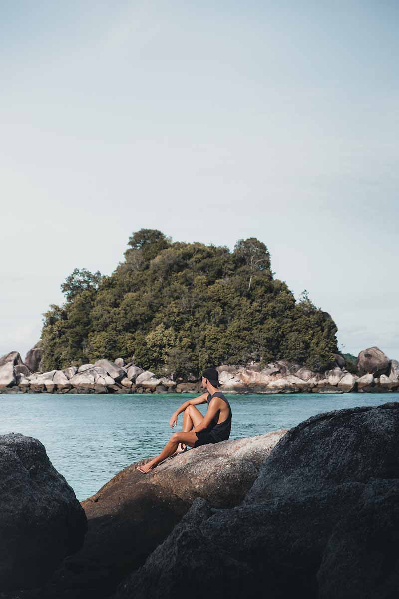 A male tourist sitting on a boulder looking out at the tiny island of Koh Usen.