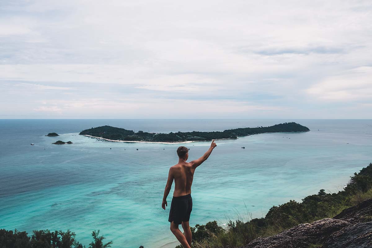 A tourist standing at Koh Lipe viewpoint with a raised right arm pointing at Koh Lipe which can clearly be seen in the distance.