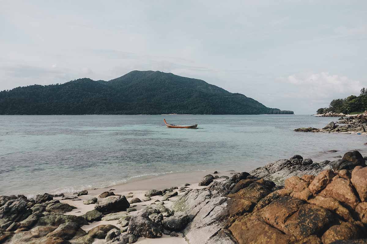 The view of Koh Adang from Pitiusas Beach with a sole long-tail boat anchored in the shallow turquoise water.
