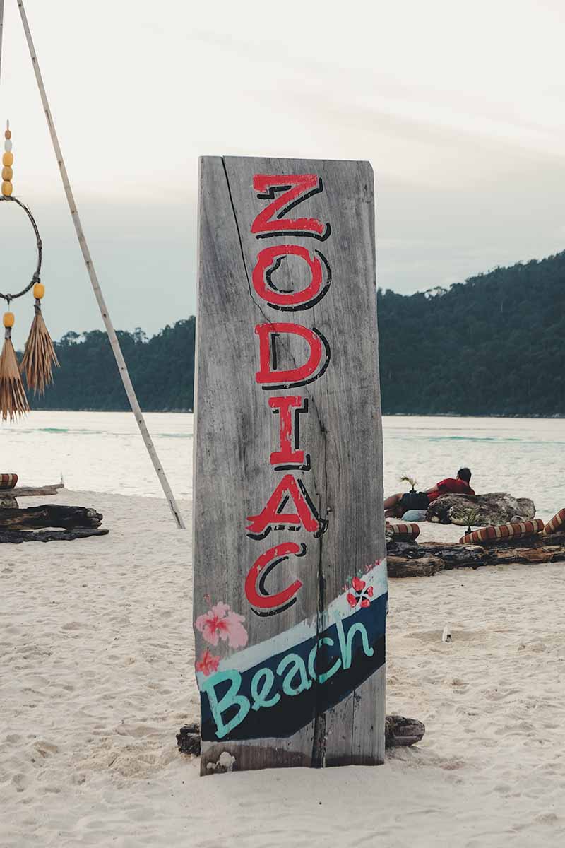 A wooden sign stuck in the sand of Sunrise Beach advertising Zodiac Beach Bar which says Zodiac Beach.