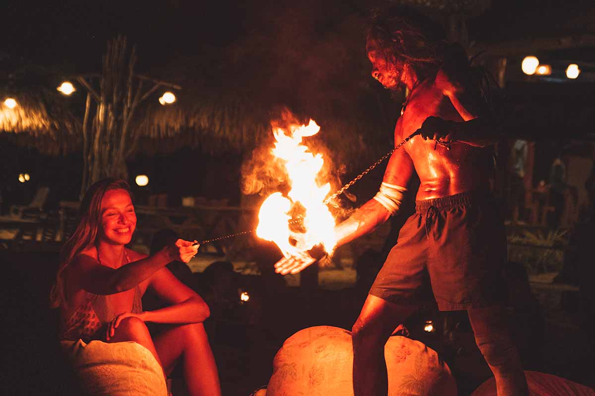 A female tourist sitting and smiling while holding one end of a fire performer's fireball chain as he holds the other end.