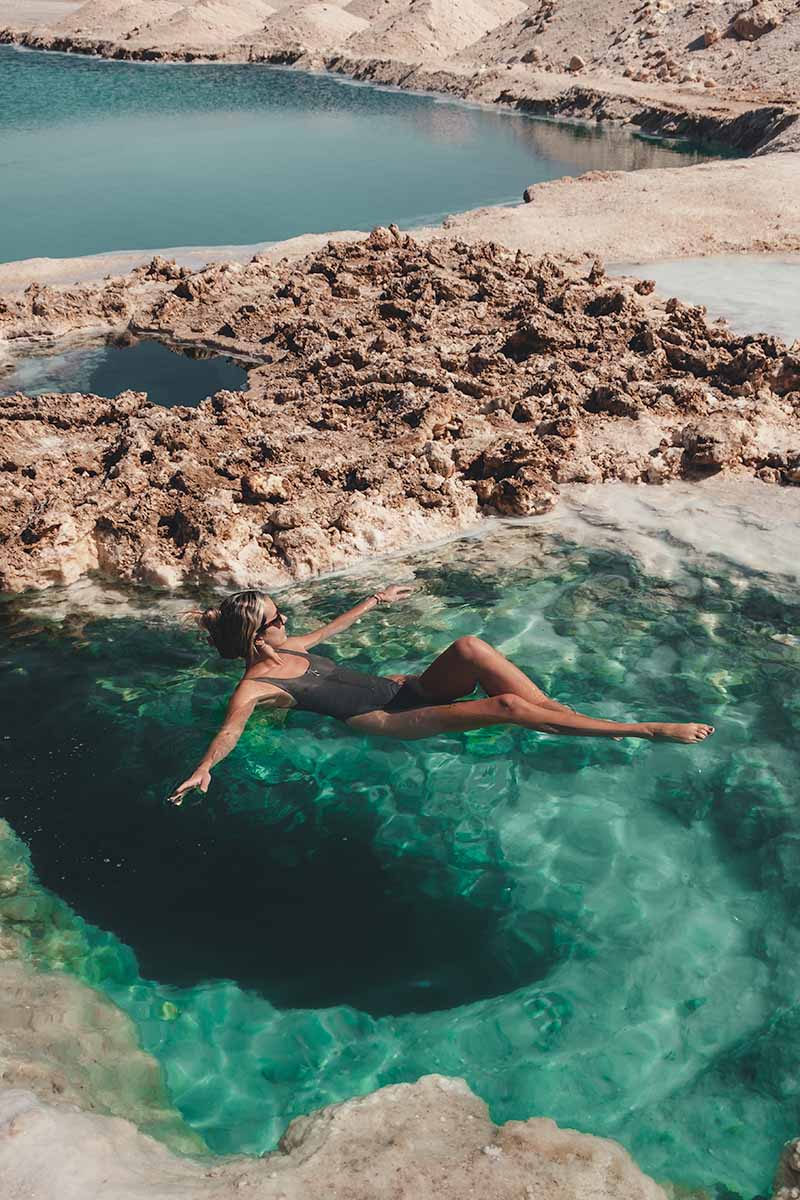 A woman floating with outstretched arms in a small emerald green salt pool within the Siwa Salt Mine.