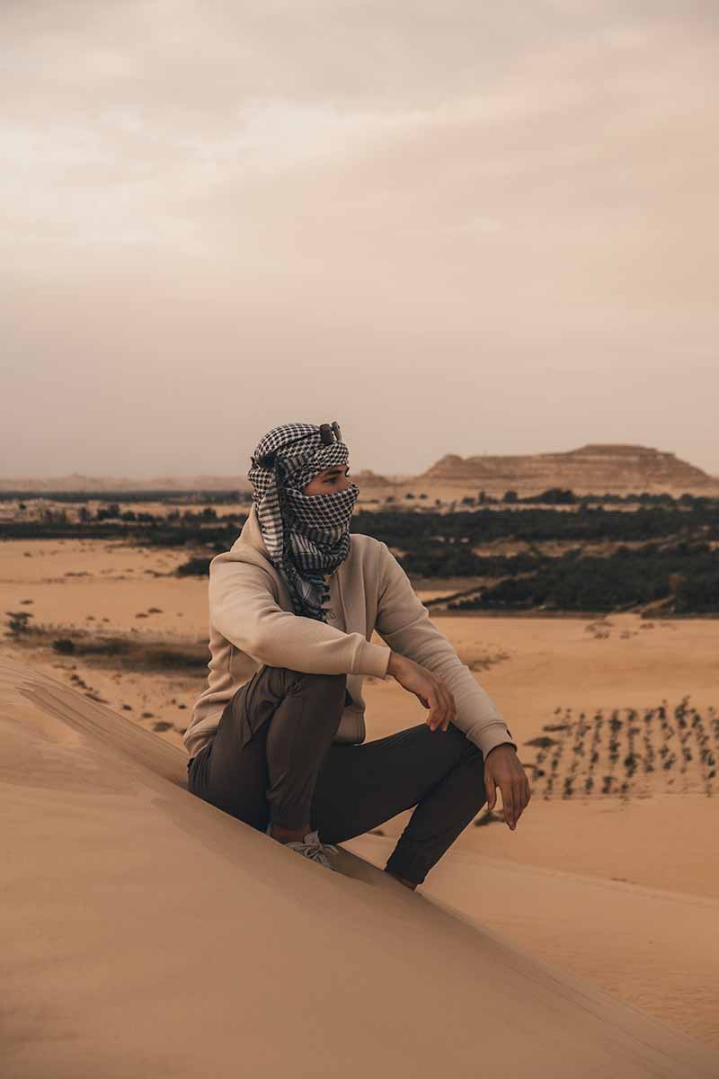 A man wearing a Keffiyeh headscarf sitting at the top of a sand dune on the outskirts of Siwa Oasis.
