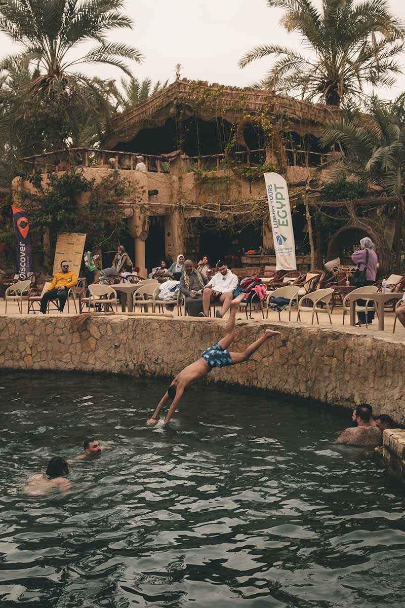 A man diving into Cleopatra's Pool while a couple of people who are sitting around the pool watch.