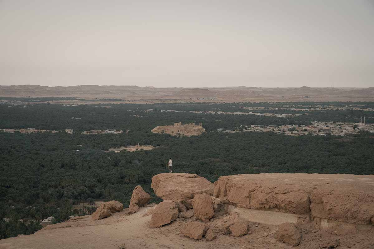 A man standing on one of the large rocks that sit at the top of Dakrur Mountain and looking out over the sweeping view of Siwa Oasis.