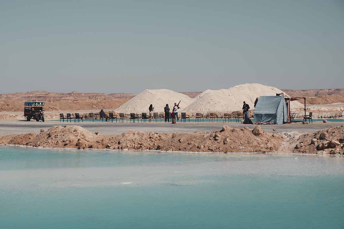 Four people standing next to a large salt pool which is line with chairs, a small tent, tuk-tuk and two large mounds of salt.