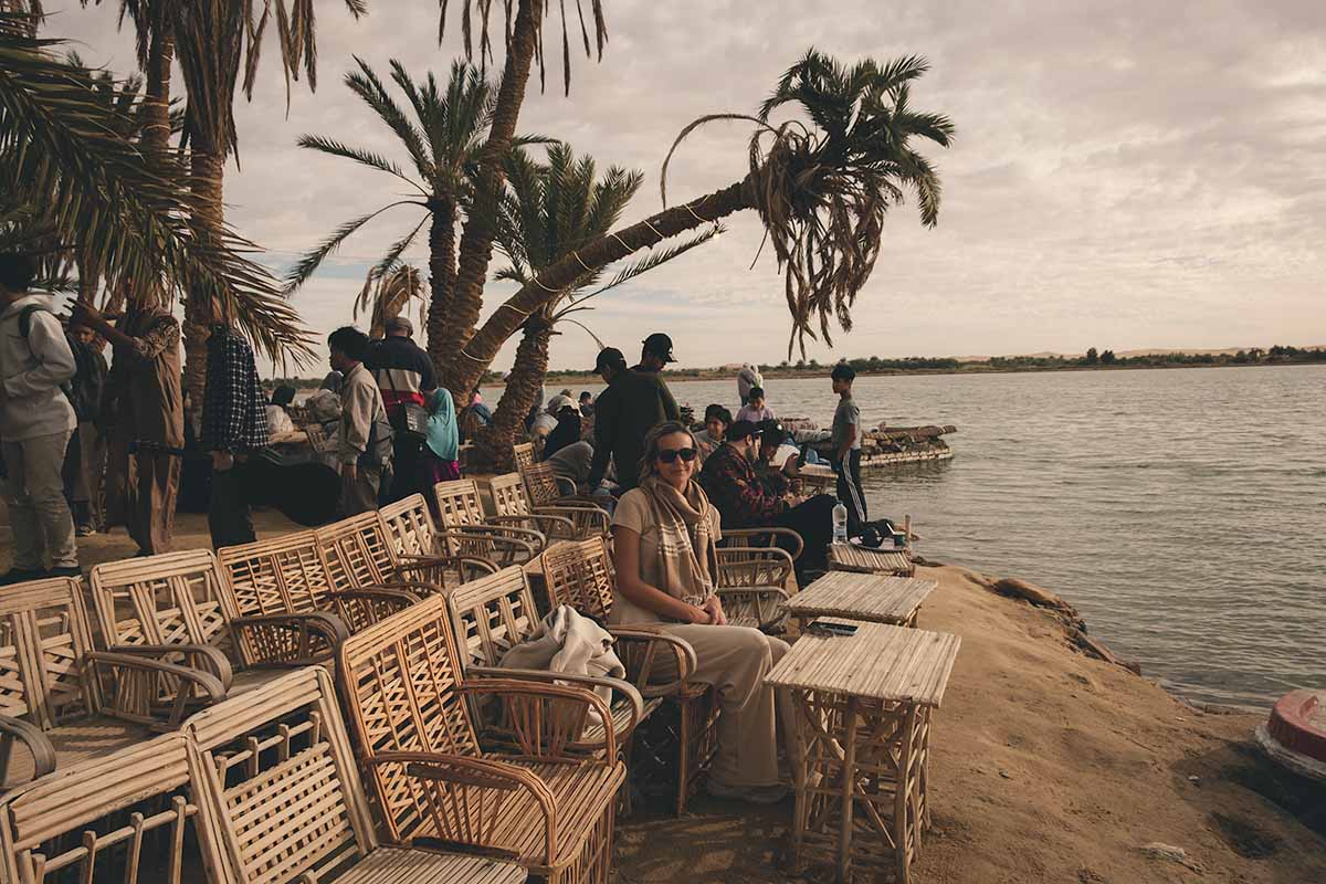 A woman sitting in the front row of lined seats next to the water of Siwa Lake who is surrounded by a large gathering of people and date palm trees.