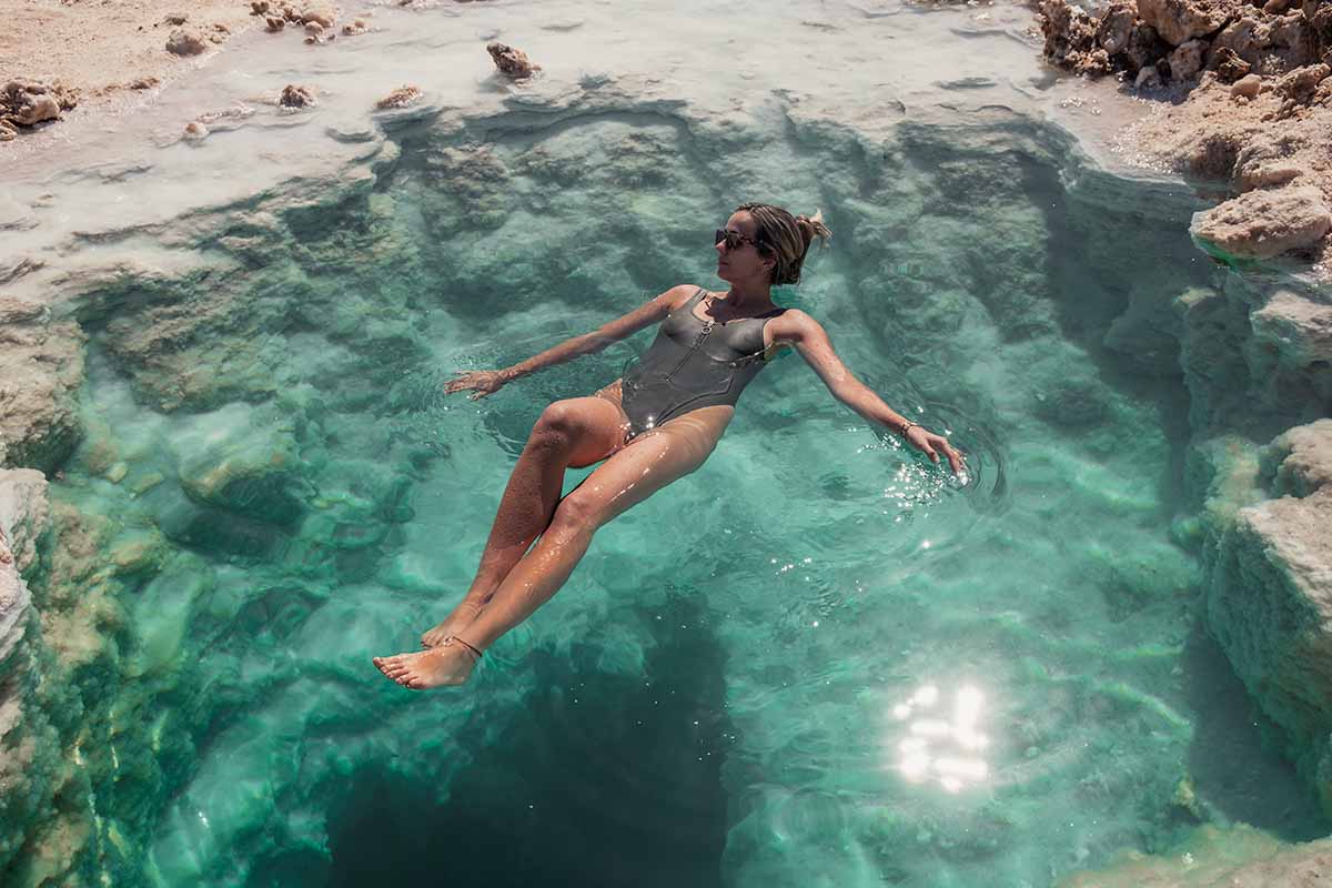 A woman in a swimming costume floating, seemingly effortlessly, in a small, turquoise salt pool that is lined with white rock salt.