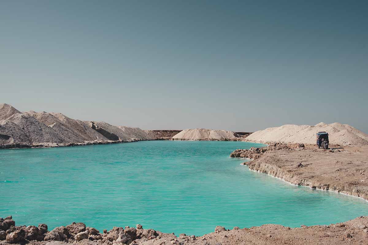 A large, rectangular turquoise salt lake surrounded by large mounds of rock salt and a single parked tuk-tuk.