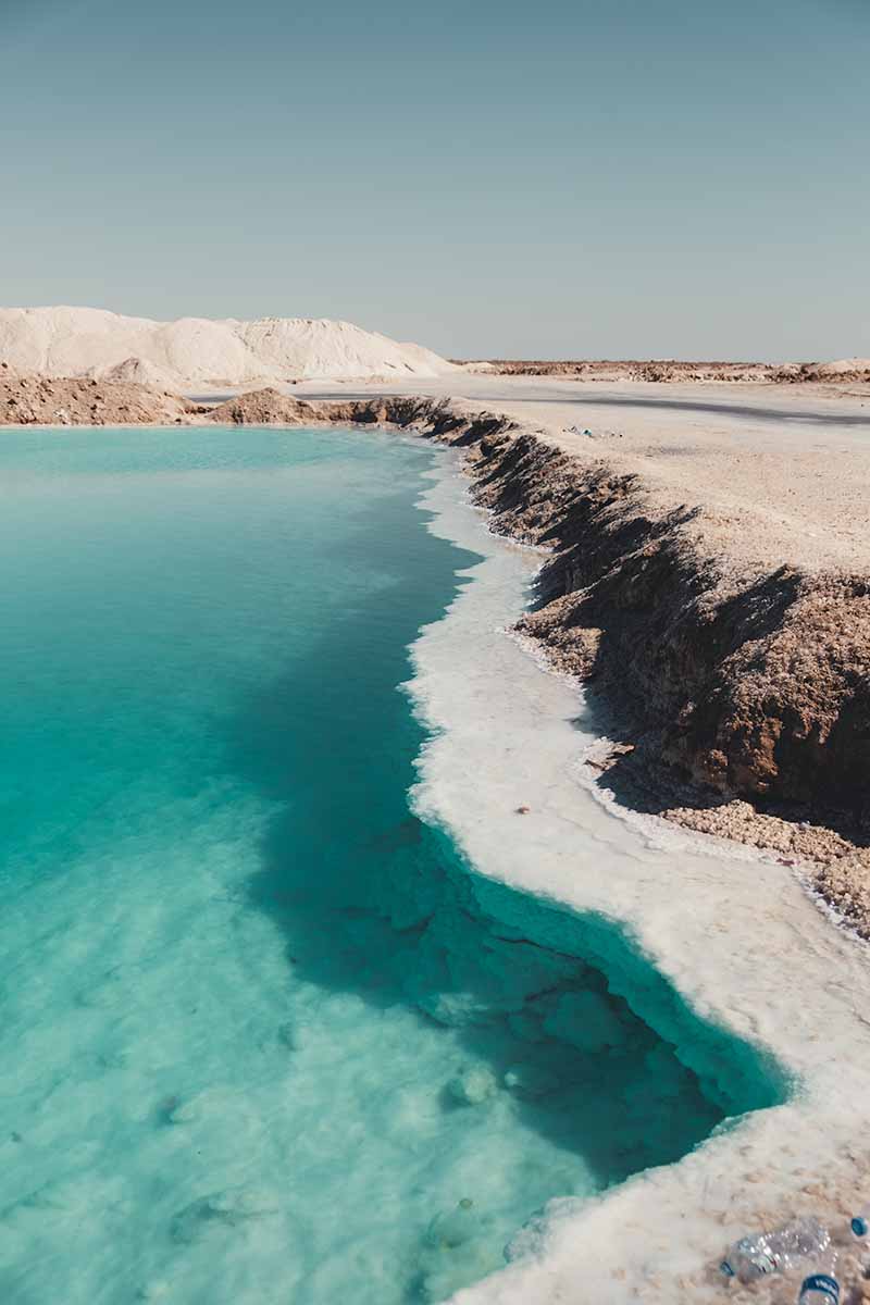 The edge of a large turquoise salt lake which is lined by white, hardened rock salt crystals.