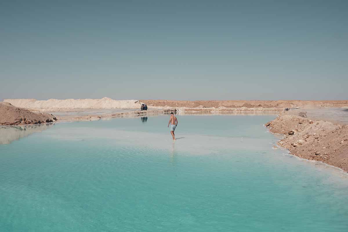 A man walking along a shallow strip in the centre of a large teal salt lake.