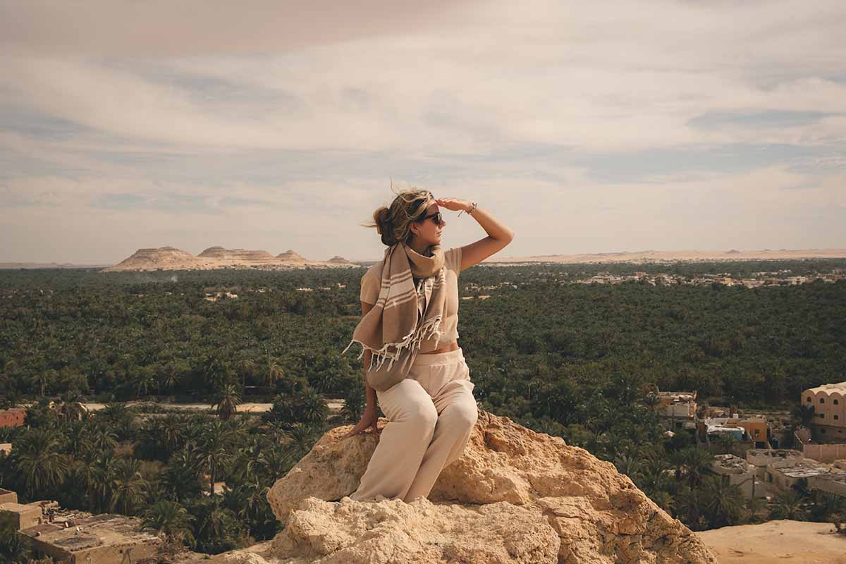 A woman sitting at the top of the Mountain of the Dead overlooking a sea of palms trees and the distant Dakrur Mountain.