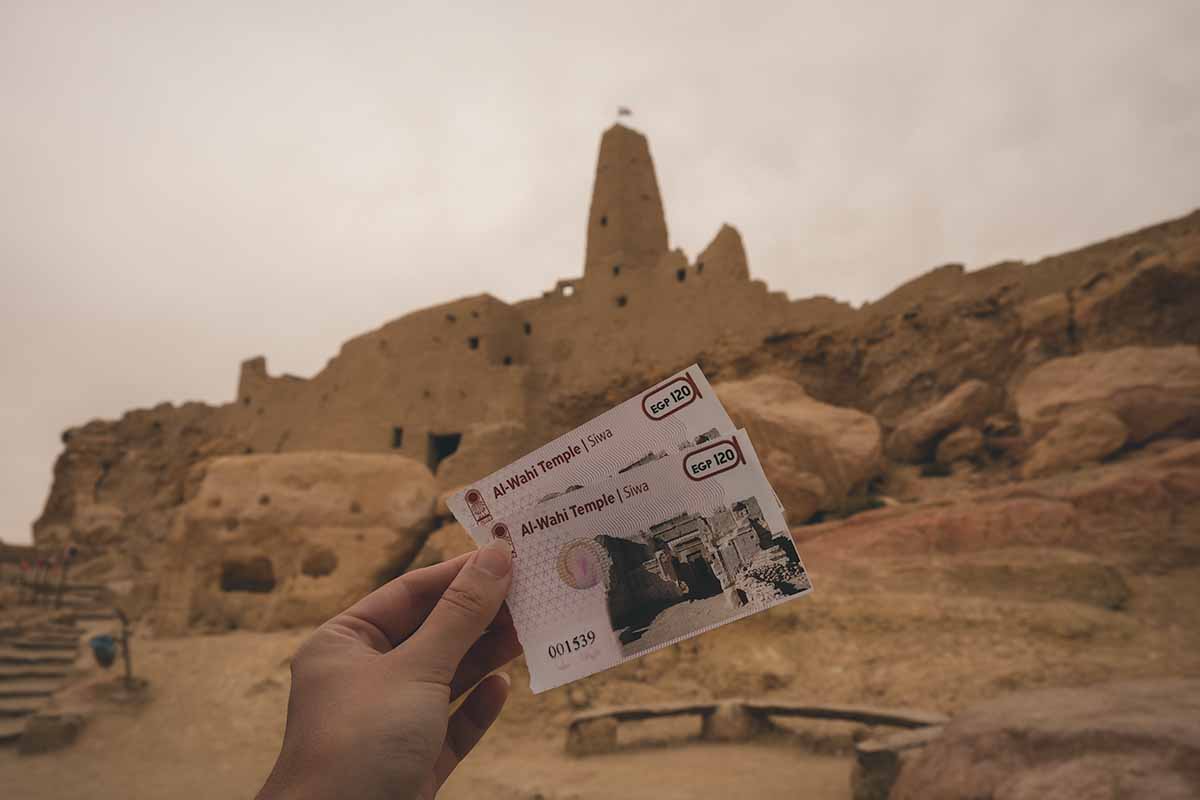 Two Al Wahi Temple Siwa entrance tickets being held up in front of the historical site.