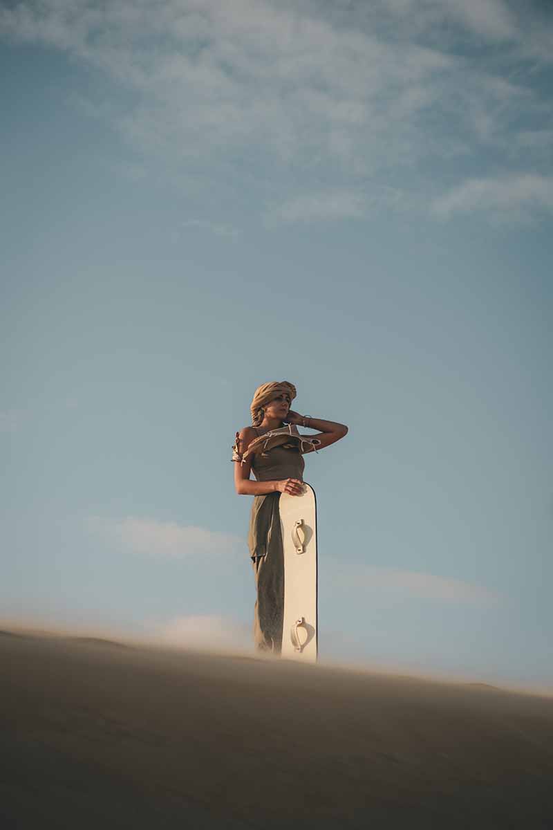 A woman standing on top of a sand dune with one hand holding her headscarf and the other a sand board.