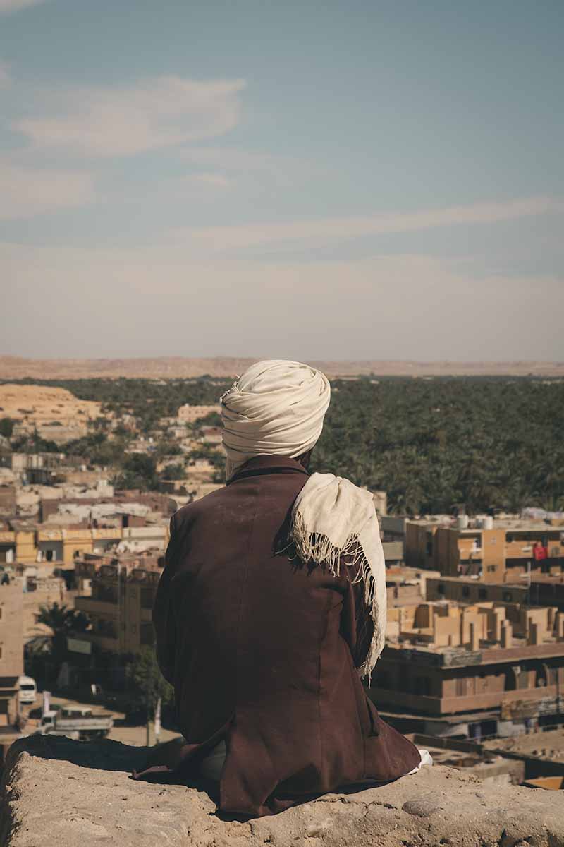 The back of a man wearing a jacket and Berber headscarf who is sitting at the top of Shali Fortress looking out over Siwa Oasis.