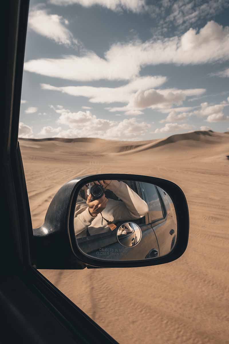 A person taking a photograph of their reflection in a jeep wing mirror from the passenger seat while the vehicle drives through the desert landscape.