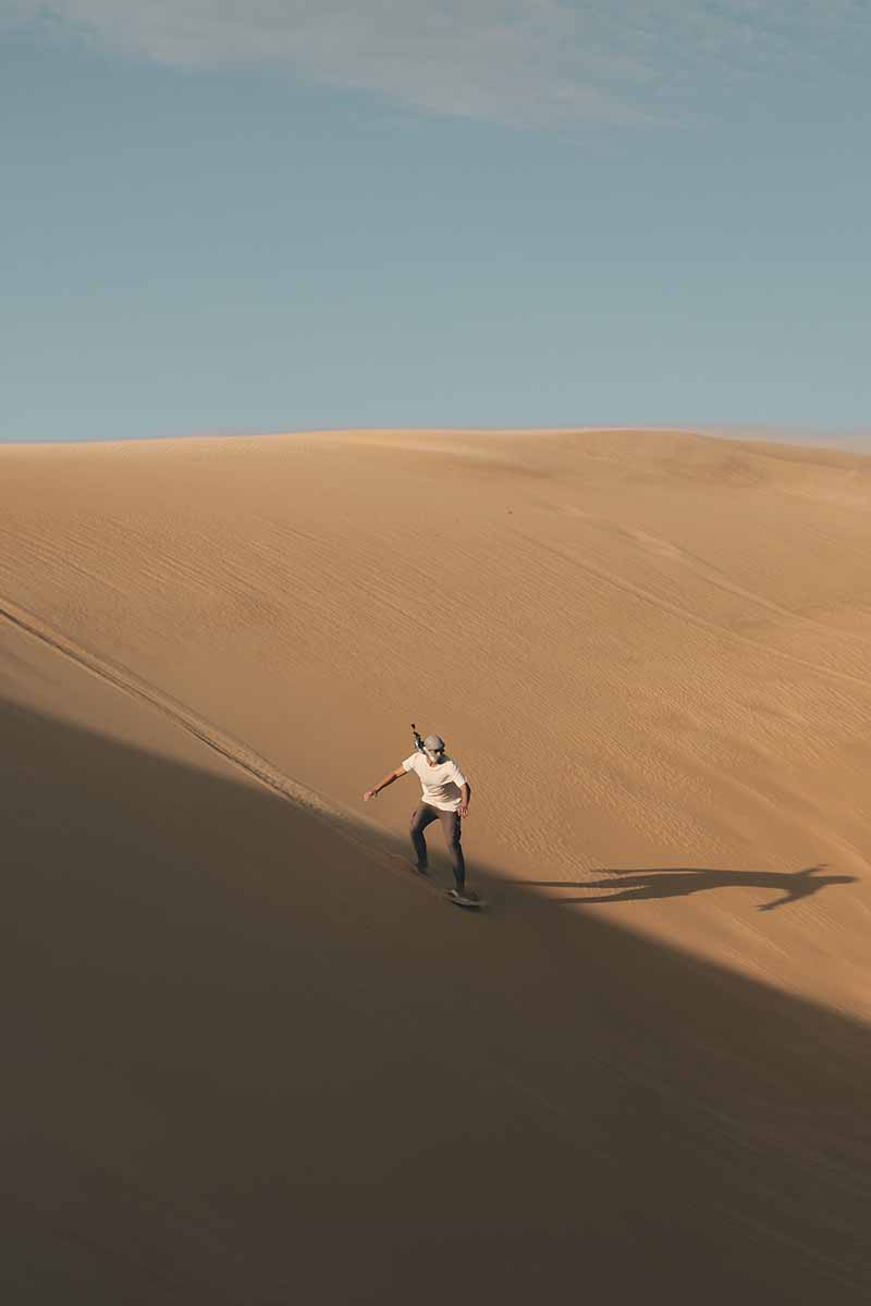 A person sand boarding down a large orange sand dune that is partly in shadow.