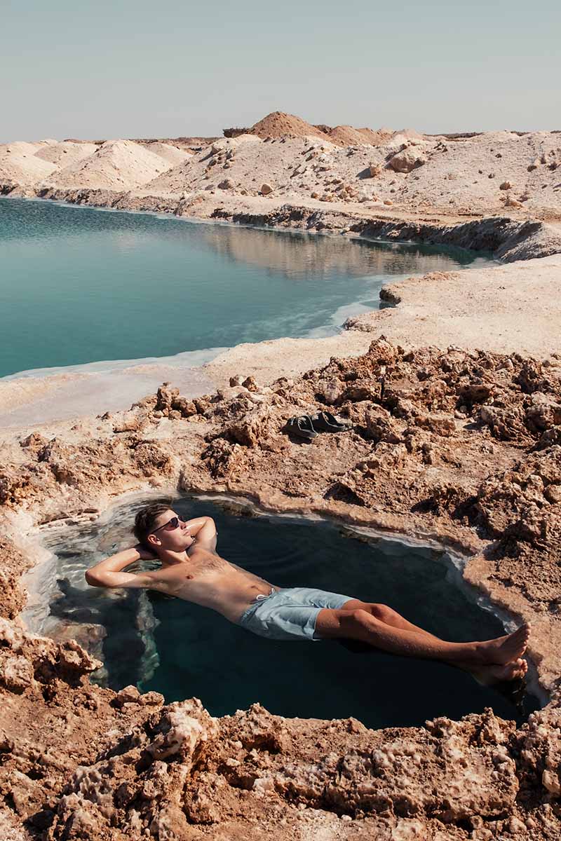 A man in swimwear floating in a dark, tiny salt pool surrounded by mounds of rock salt.