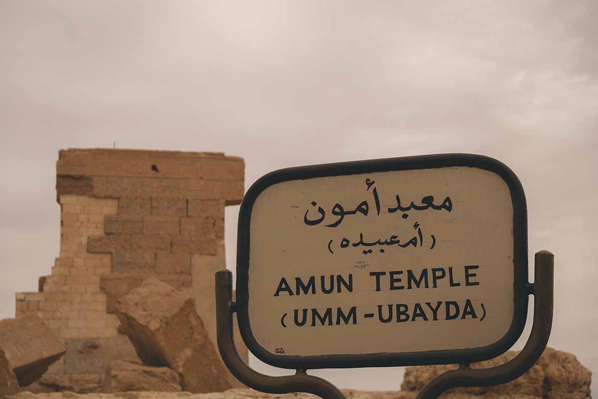 A sign readin Amun Temple (Umm - Ubayda) in English and Arabic with the remains of the temple in the background.