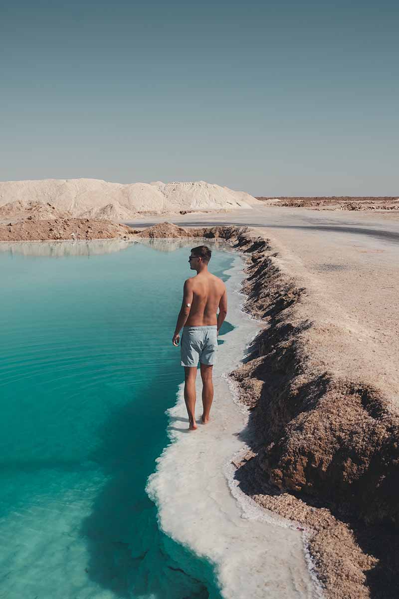 A man looking out over a large teal salt pool while walking along the crystallised edge.