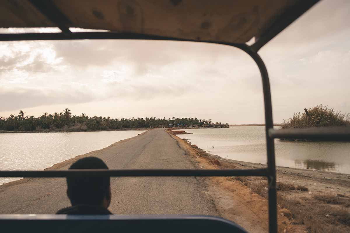 A narrow strip of raised road surrounded by Fatnas Lake leading to the palm tree-covered Fatnas Island.