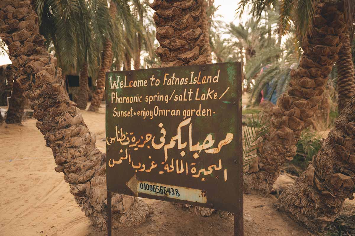 A sign at the entrance to Fatnas Island that reads "Welcome to Fatnas Island Pharaonic spring/salt lake/ Sunset. Enjoy Omran garden" with an Arabic translation and contact number underneath.