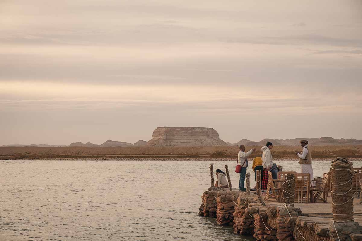 A handful of people at the end of a jetty on Fatnas Lake with giant desert rock plateaus in the distance.