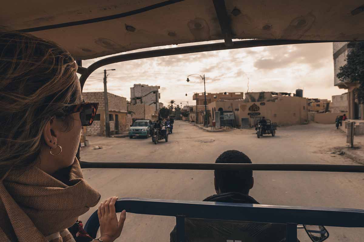 A woman sitting in the back of a tuk-tuk that is being driven through the dusty streets of Siwa Oasis.