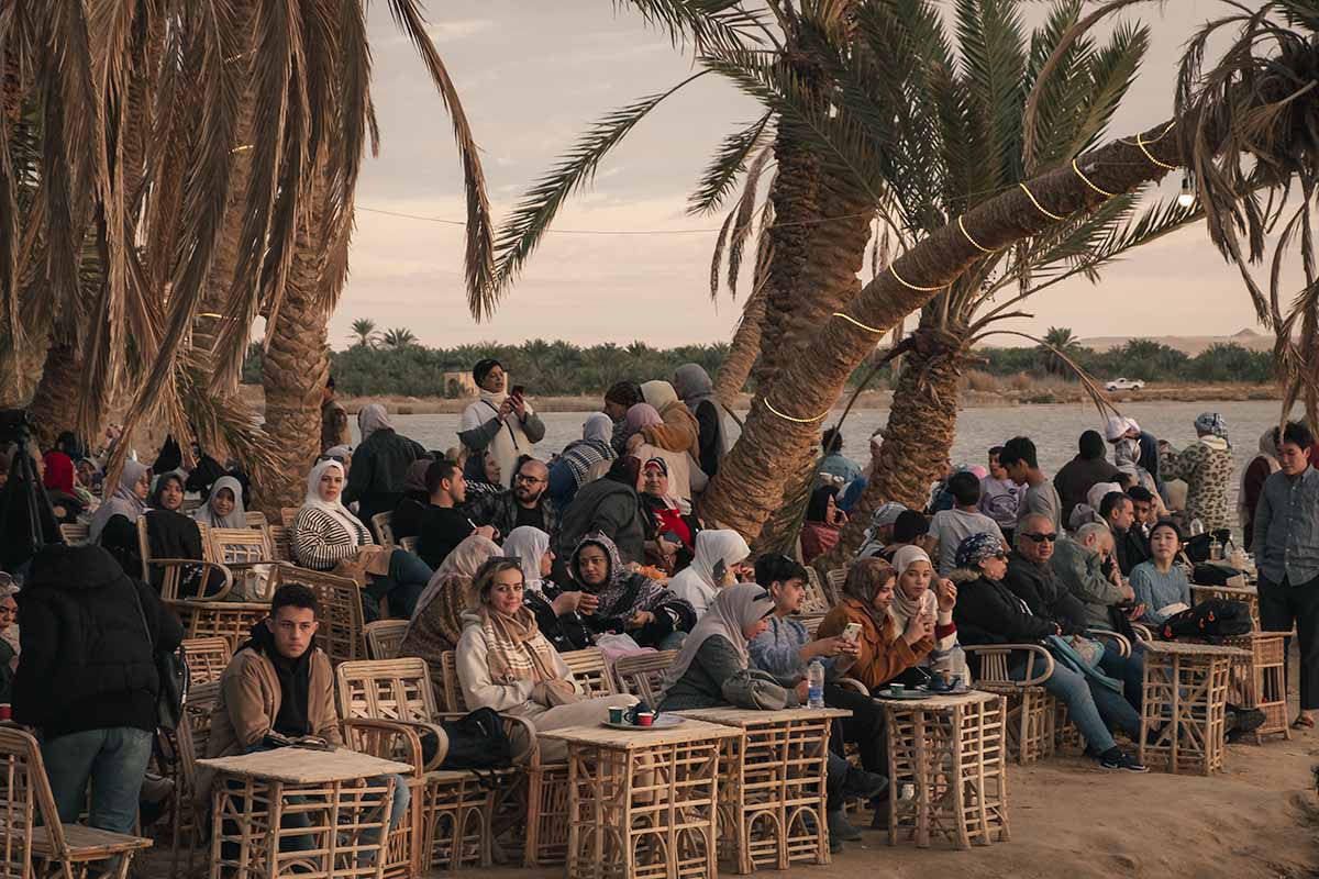 Several rows of people sitting on wooden chairs amongst date palm trees.