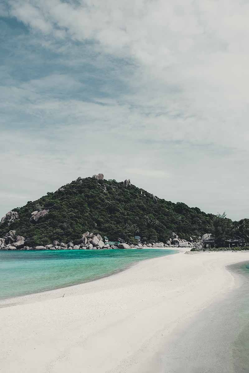 A narrow strip of white sand with water on either side leading to a rocky, jungle-like islet.