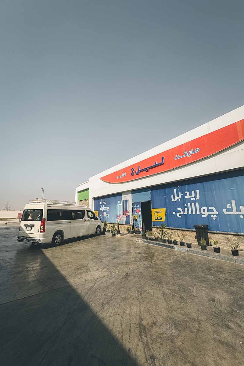 A white minivan parked outside a service station which is displaying Arabic adverts in the windows.