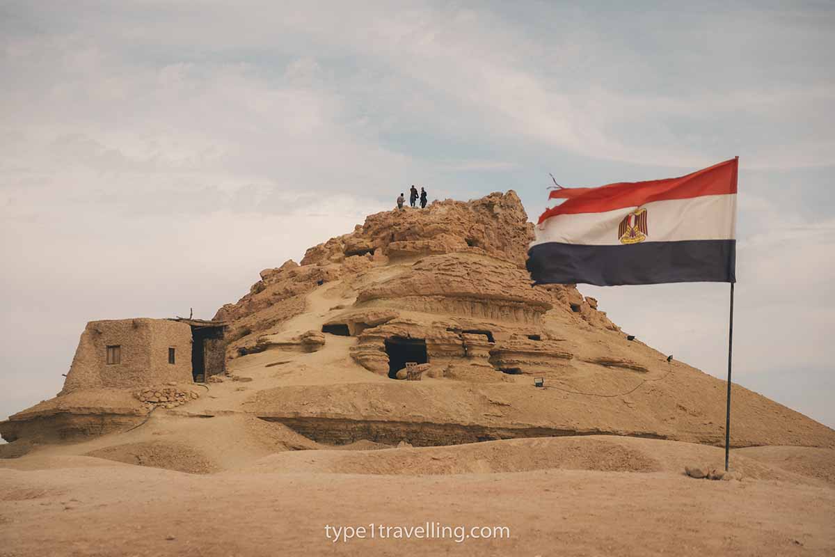 An Egyptian flag flying at the bottom of Gabal al Mawta while a group of tourists stand at the top.